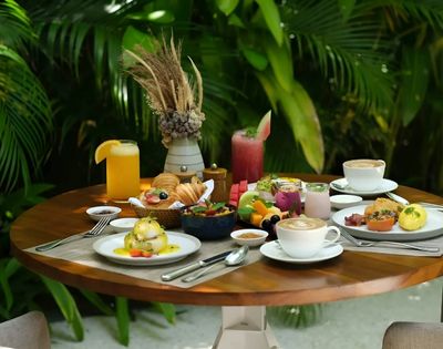 Alt text: A round wooden table set for a tropical brunch featuring pastries, fruit, poached eggs, and various drinks, set against lush green foliage.