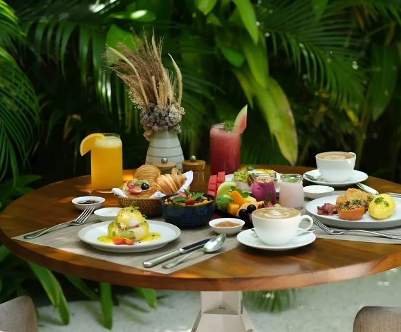 Alt text: A round wooden table set for a tropical brunch featuring pastries, fruit, poached eggs, and various drinks, set against lush green foliage.