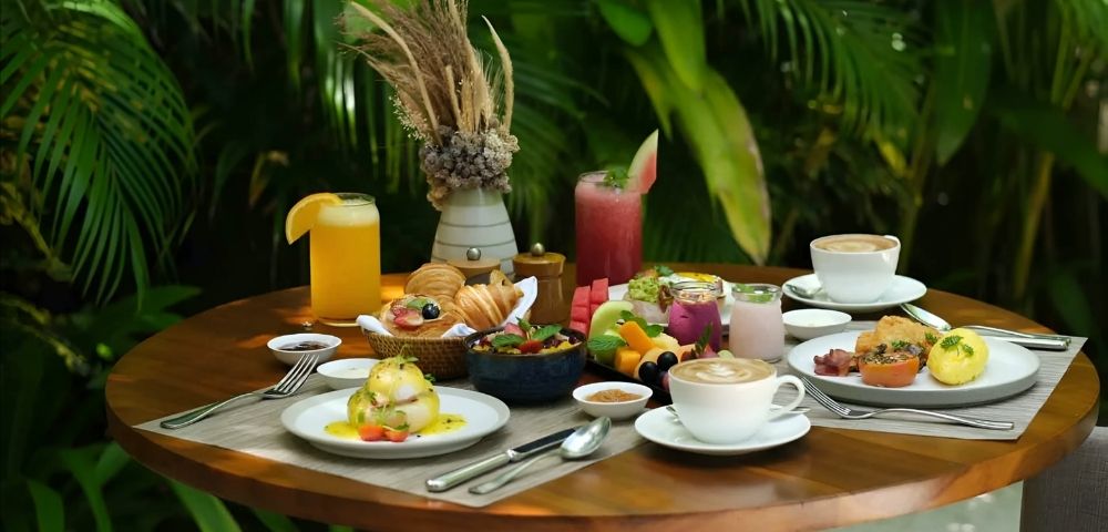 Alt text: A round wooden table set for a tropical brunch featuring pastries, fruit, poached eggs, and various drinks, set against lush green foliage.