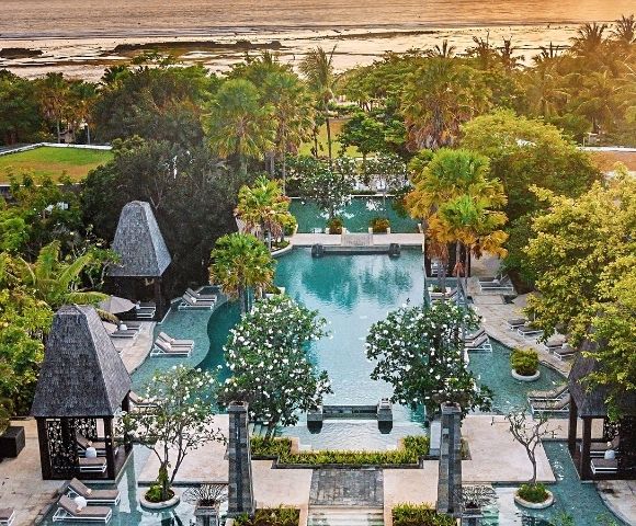 Aerial view of a luxurious tropical resort with a central pool surrounded by lush greenery and palm trees, leading to a serene ocean at sunset.