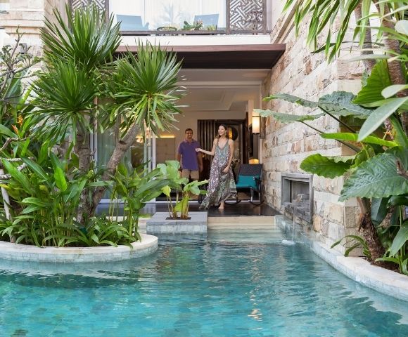 A couple stands by a luxurious stone villa with a clear turquoise pool, surrounded by lush green plants, creating a serene and tropical atmosphere.