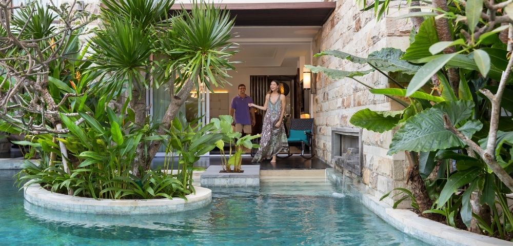 A couple stands by a luxurious stone villa with a clear turquoise pool, surrounded by lush green plants, creating a serene and tropical atmosphere.