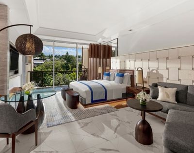 Modern hotel room with a large bed, blue accents, and a glass table. Floor-to-ceiling windows reveal lush greenery, creating a serene atmosphere.