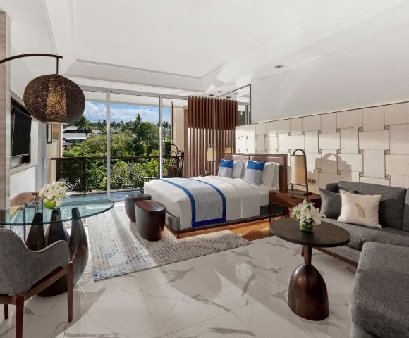 Modern hotel room with a large bed, blue accents, and a glass table. Floor-to-ceiling windows reveal lush greenery, creating a serene atmosphere.