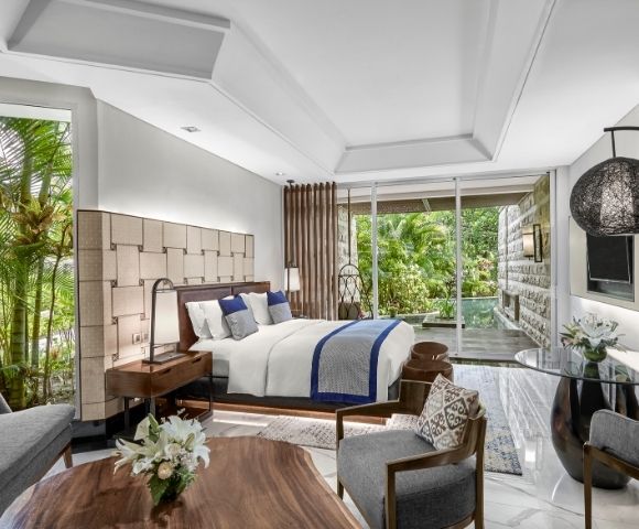 Luxurious hotel room with a plush bed, wooden furnishings, and large windows revealing a lush garden. Modern decor creates a tranquil, inviting ambiance.