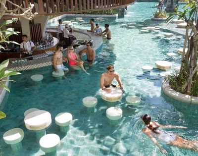 People relaxing at a swim-up bar in a tropical pool. Several are sitting on submerged stools while others swim. A bartender serves drinks, creating a lively and refreshing scene.