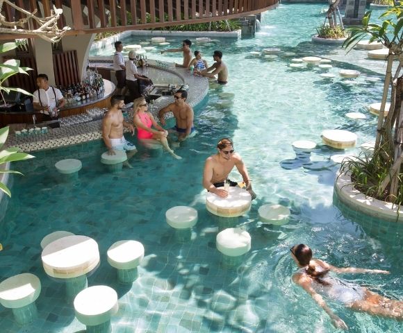 People relaxing at a swim-up bar in a tropical pool. Several are sitting on submerged stools while others swim. A bartender serves drinks, creating a lively and refreshing scene.