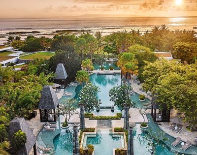 Aerial view of a tropical resort at sunset. Elegant pools are surrounded by lush greenery and decorative stone structures. The ocean glimmers in the background.