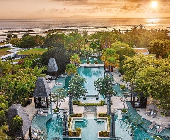 Aerial view of a tropical resort at sunset. Elegant pools are surrounded by lush greenery and decorative stone structures. The ocean glimmers in the background.