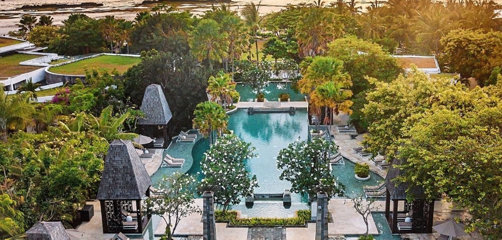 Aerial view of a tropical resort at sunset. Elegant pools are surrounded by lush greenery and decorative stone structures. The ocean glimmers in the background.