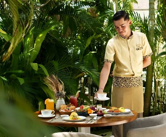 A waiter in a beige uniform places a dish on a table with a tropical breakfast spread, surrounded by lush green plants, creating a serene and inviting atmosphere.