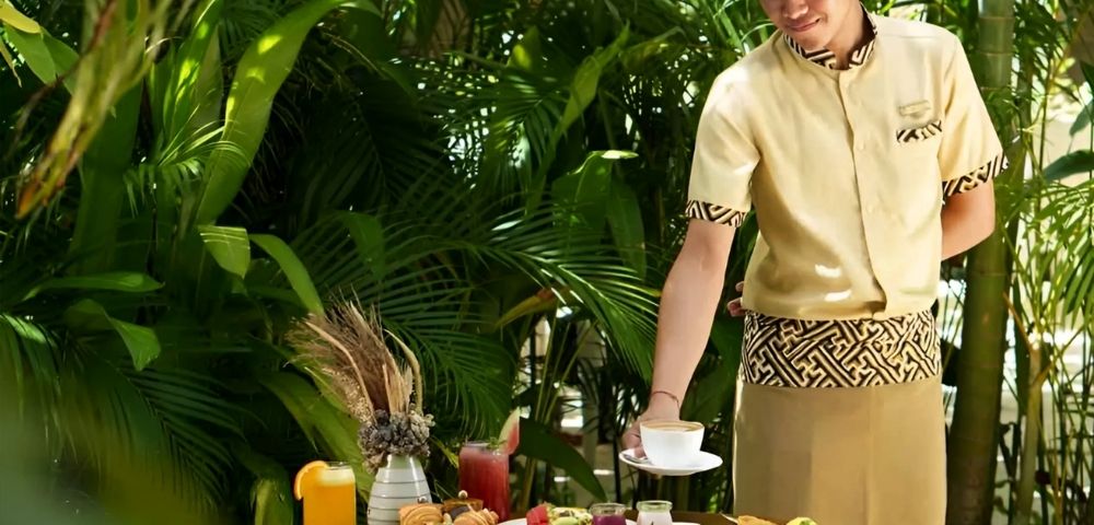 A waiter in a beige uniform places a dish on a table with a tropical breakfast spread, surrounded by lush green plants, creating a serene and inviting atmosphere.