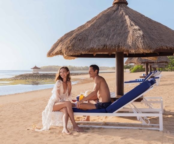 A man and woman relax on beach loungers under a straw hut, smiling amidst a sunny seaside setting with a serene ocean backdrop.