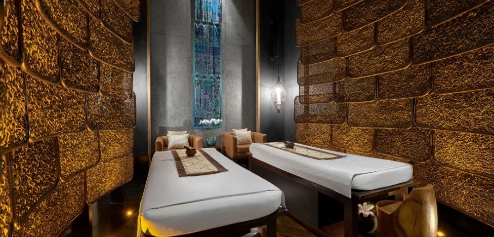 Luxurious spa room with two massage tables, earthy tones, dim lighting, and textured walls. A serene, calming atmosphere is created by ambient light.