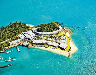 Aerial view of a lush, green island resort surrounded by turquoise water. White buildings, a marina, and sandy beaches convey a tranquil, tropical vibe.