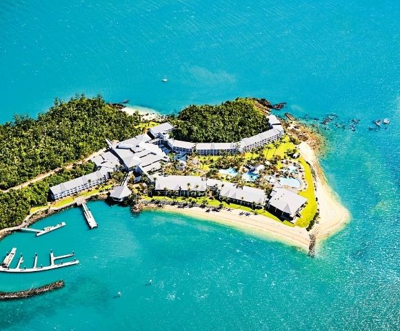 Aerial view of a lush, green island resort surrounded by turquoise water. White buildings, a marina, and sandy beaches convey a tranquil, tropical vibe.