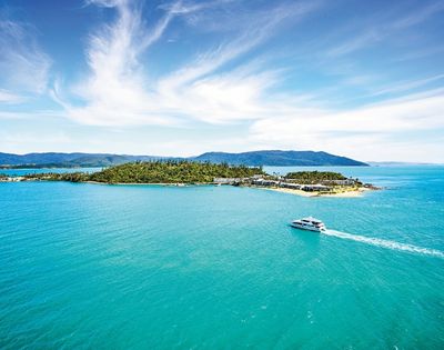 A serene island surrounded by turquoise waters under a clear blue sky. A boat glides across the water, leaving a white trail, suggesting leisure and tranquility.