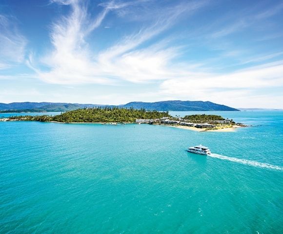 A serene island surrounded by turquoise waters under a clear blue sky. A boat glides across the water, leaving a white trail, suggesting leisure and tranquility.