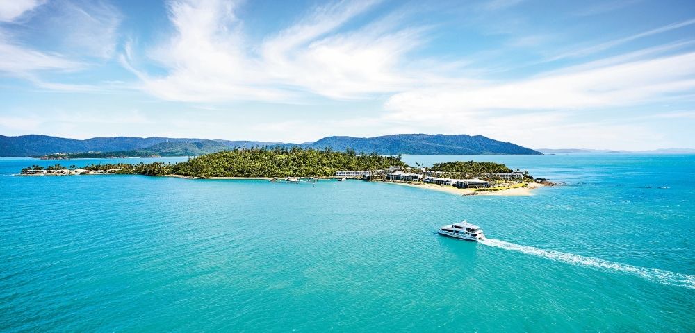 A serene island surrounded by turquoise waters under a clear blue sky. A boat glides across the water, leaving a white trail, suggesting leisure and tranquility.