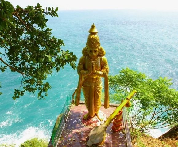 Golden statue on a cliff overlooks vast blue ocean, surrounded by green trees. The serene setting conveys tranquility and connection with nature.