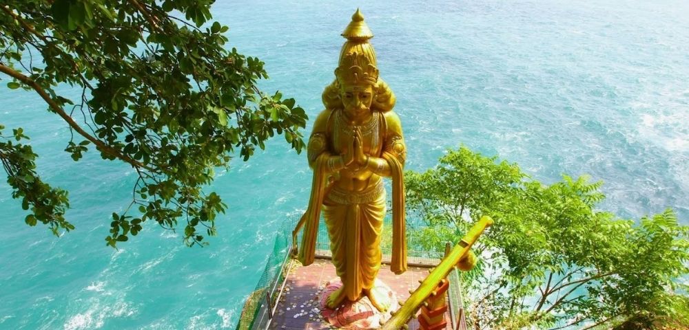 Golden statue on a cliff overlooks vast blue ocean, surrounded by green trees. The serene setting conveys tranquility and connection with nature.
