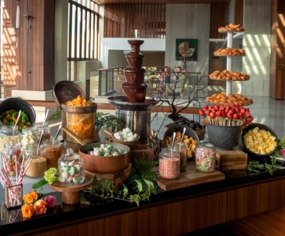 A chocolate fountain surrounded by assorted treats, including fruits, marshmallows, and pastries, creating a lavish, inviting dessert display.