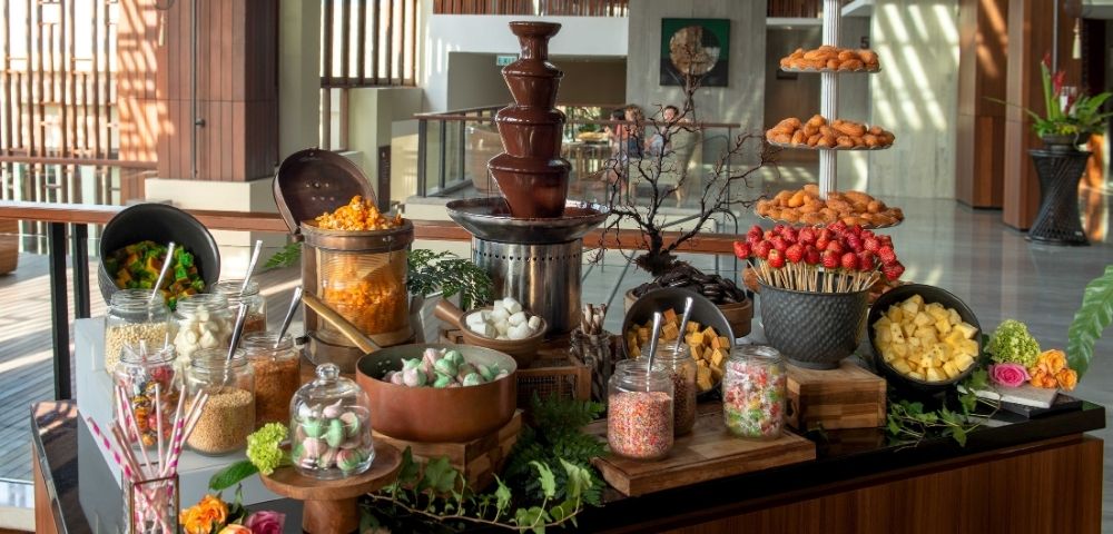 A chocolate fountain surrounded by assorted treats, including fruits, marshmallows, and pastries, creating a lavish, inviting dessert display.