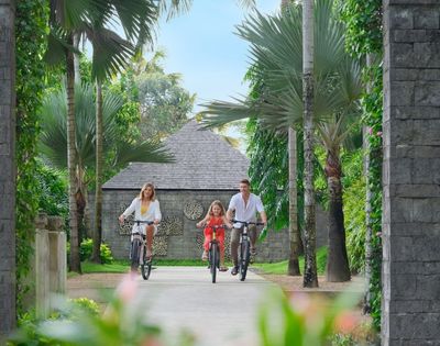 A family of three, joyfully cycling along a lush, tropical path. Tall palm trees line the way, with a stone house and vibrant greenery in the background.