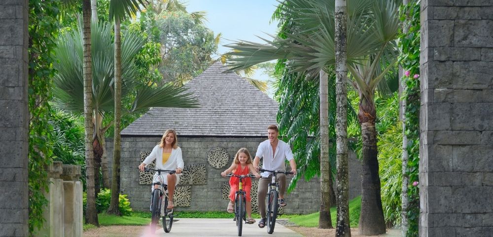 A family of three, joyfully cycling along a lush, tropical path. Tall palm trees line the way, with a stone house and vibrant greenery in the background.