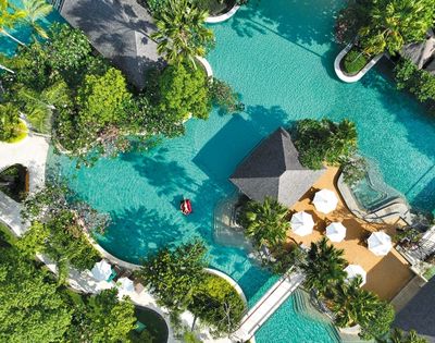 Aerial view of a tropical resort pool surrounded by lush greenery. A person relaxes on a red float in turquoise water near cabanas and white umbrellas.