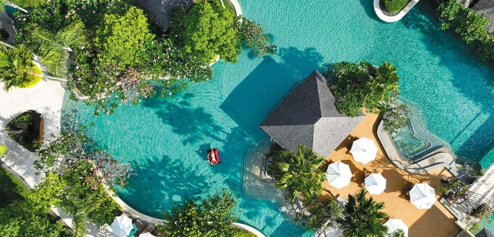 Aerial view of a tropical resort pool surrounded by lush greenery. A person relaxes on a red float in turquoise water near cabanas and white umbrellas.