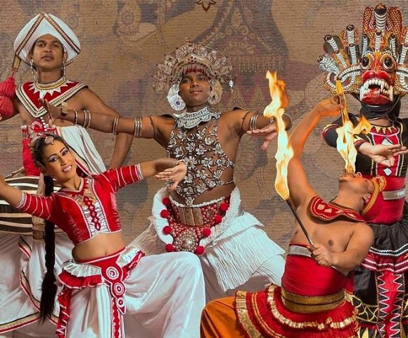 A vibrant traditional Sri Lankan cultural dance performance featuring performers in ornate costumes, including colorful garments, decorative headdresses, and expressive hand and body movements, set against a textured backdrop with cultural motifs.