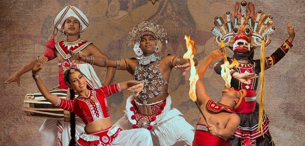 A vibrant traditional Sri Lankan cultural dance performance featuring performers in ornate costumes, including colorful garments, decorative headdresses, and expressive hand and body movements, set against a textured backdrop with cultural motifs.