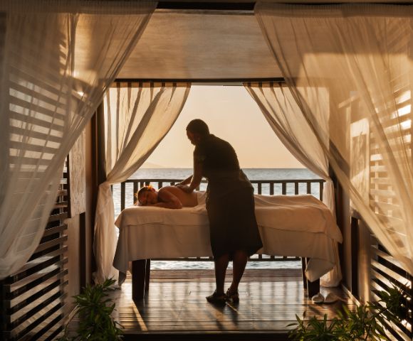 A person receives a relaxing massage on a bed in an open-air cabana with sheer curtains. The serene setting overlooks a calm ocean at sunset.