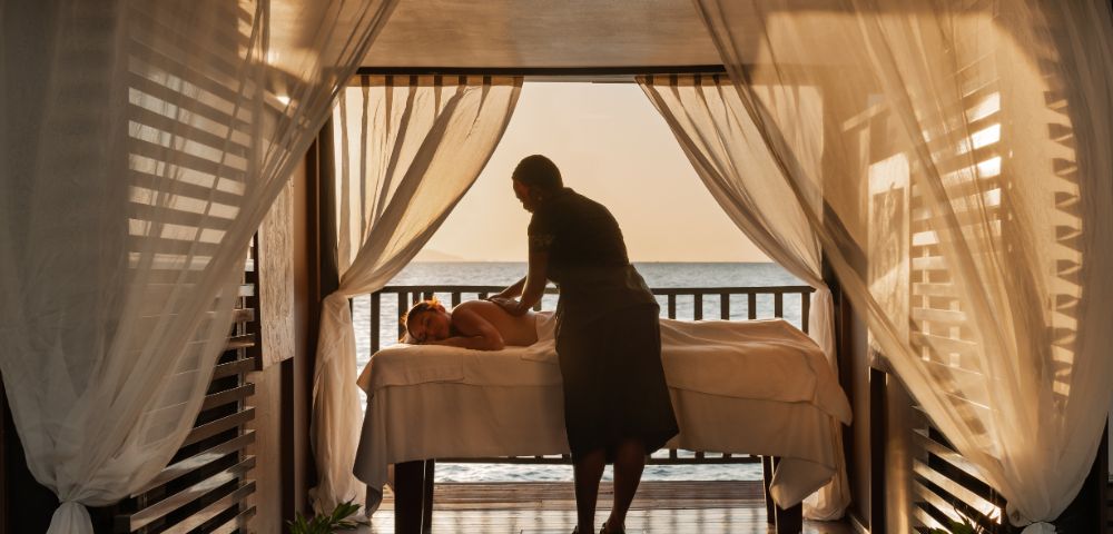 A person receives a relaxing massage on a bed in an open-air cabana with sheer curtains. The serene setting overlooks a calm ocean at sunset.