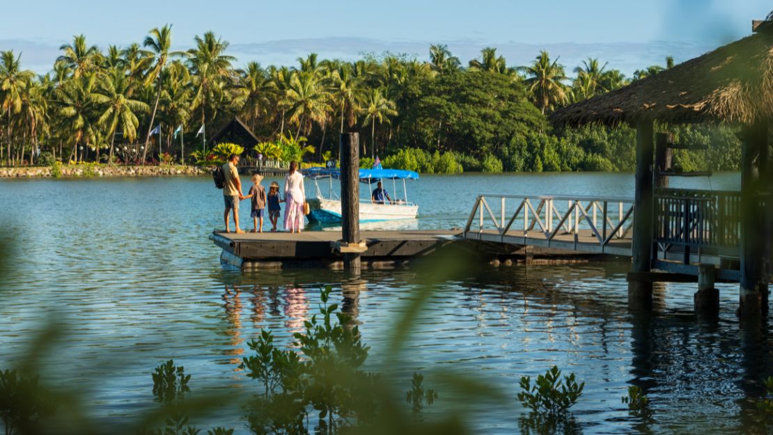 People stand on a wooden dock by a tranquil, palm-fringed lagoon. A small boat with a blue canopy is moored nearby. The scene is peaceful and tropical.