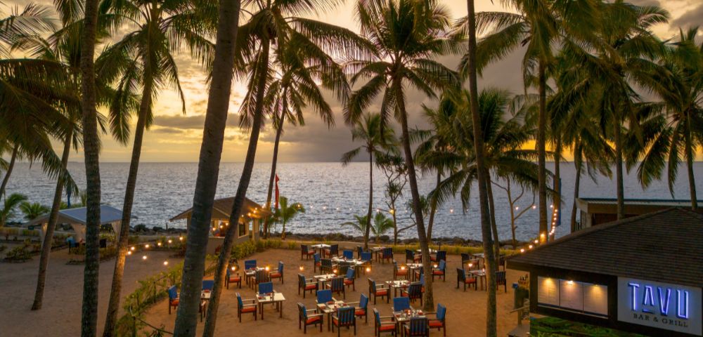 Outdoor beach restaurant at sunset, framed by tall palm trees. Tables are set with blue chairs, creating a tranquil and inviting seaside atmosphere.