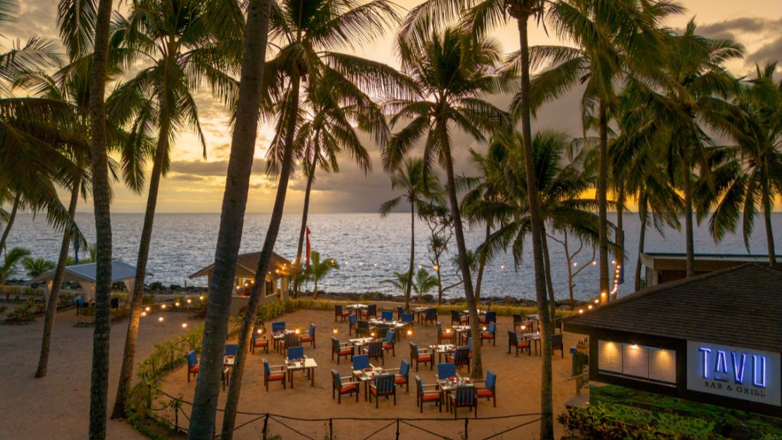 Outdoor beach restaurant at sunset, framed by tall palm trees. Tables are set with blue chairs, creating a tranquil and inviting seaside atmosphere.