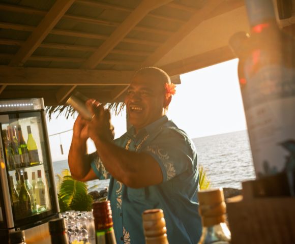 A cheerful bartender in a blue shirt shakes a cocktail shaker at a beachside bar during sunset, with bottles and a fridge of drinks nearby.