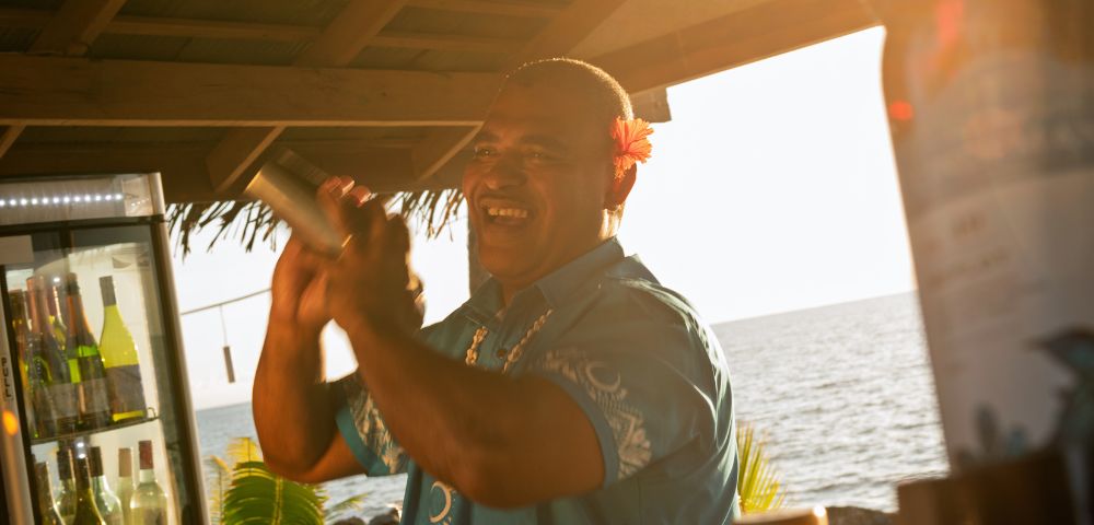 A cheerful bartender in a blue shirt shakes a cocktail shaker at a beachside bar during sunset, with bottles and a fridge of drinks nearby.
