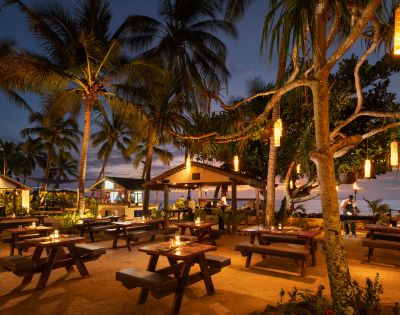 Tropical beach restaurant at dusk with wooden tables, flickering candles, and hanging lanterns. Palm trees and sea create a serene, inviting ambiance.