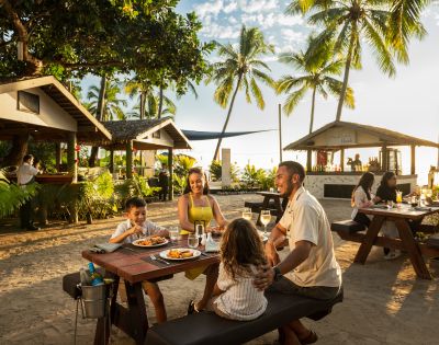 Family dining outdoors at a tropical beachside restaurant. Palm trees, sunlight, and a relaxed atmosphere set the scene. Smiles and warmth abound.