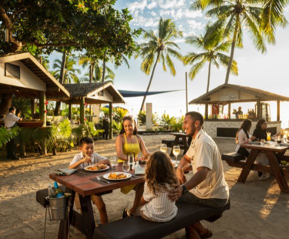 Family dining outdoors at a tropical beachside restaurant. Palm trees, sunlight, and a relaxed atmosphere set the scene. Smiles and warmth abound.