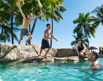 Two boys jump into a tropical pool while a man and woman watch, surrounded by palm trees and a clear blue sky, conveying joy and relaxation.