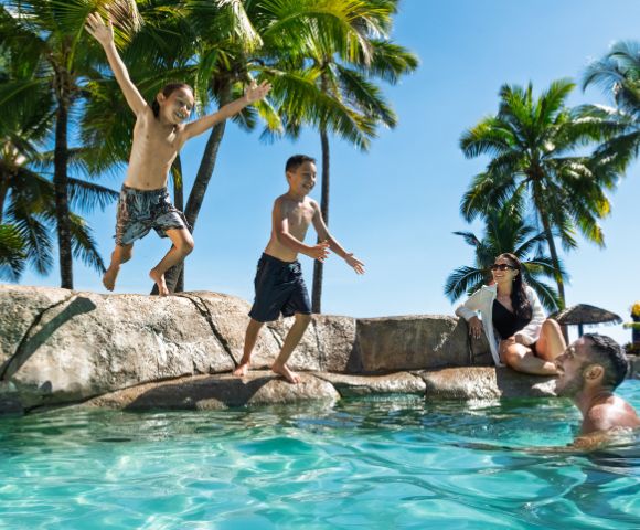 Two boys jump into a tropical pool while a man and woman watch, surrounded by palm trees and a clear blue sky, conveying joy and relaxation.