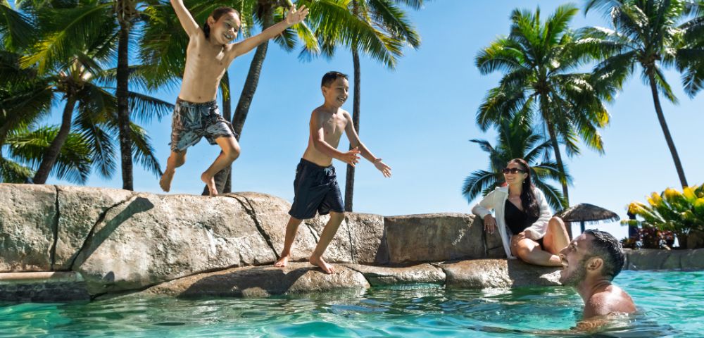 Two boys jump into a tropical pool while a man and woman watch, surrounded by palm trees and a clear blue sky, conveying joy and relaxation.