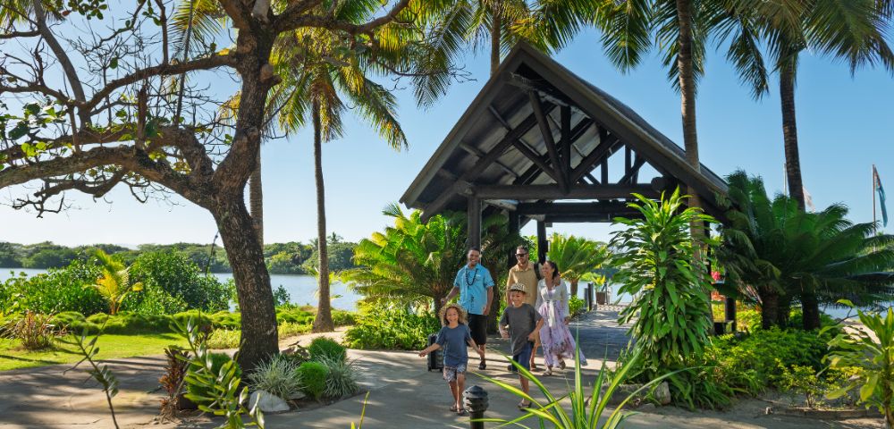 A family of five walks on a sunny path surrounded by lush tropical greenery and palm trees. A wooden pavilion and a glimpse of water are in the background.