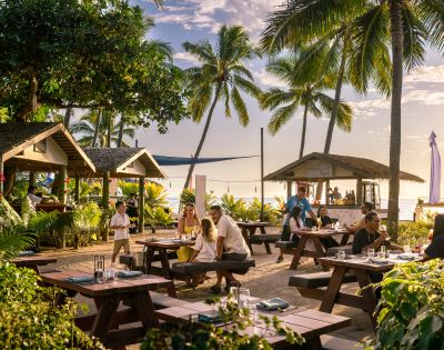 Tropical beachside café with wooden tables, lush greenery, and palm trees. Diners enjoy a relaxed meal near the ocean at sunset, creating a warm, inviting atmosphere.