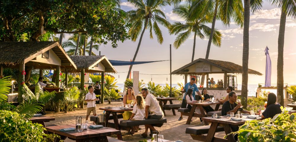 Tropical beachside café with wooden tables, lush greenery, and palm trees. Diners enjoy a relaxed meal near the ocean at sunset, creating a warm, inviting atmosphere.