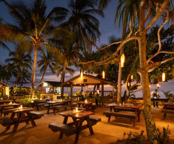 Outdoor beach restaurant at dusk with wooden tables lit by candlelight. Surrounded by palm trees and lanterns, creating a warm, inviting atmosphere.